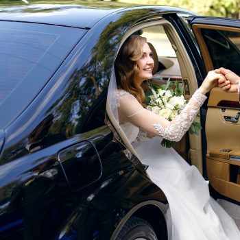 Groom helps happy young bride to get out of wedding car. Elegant bride holds wedding bouquet of white roses in her hands. The bride and groom arrived at the wedding ceremony